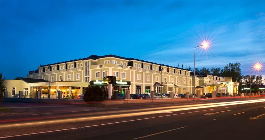 Wild Ireland - A large, well-lit hotel building with multiple windows and a flat roof sits beside a road at dusk. Cars are parked outside, and light trails from passing vehicles are visible in the foreground.