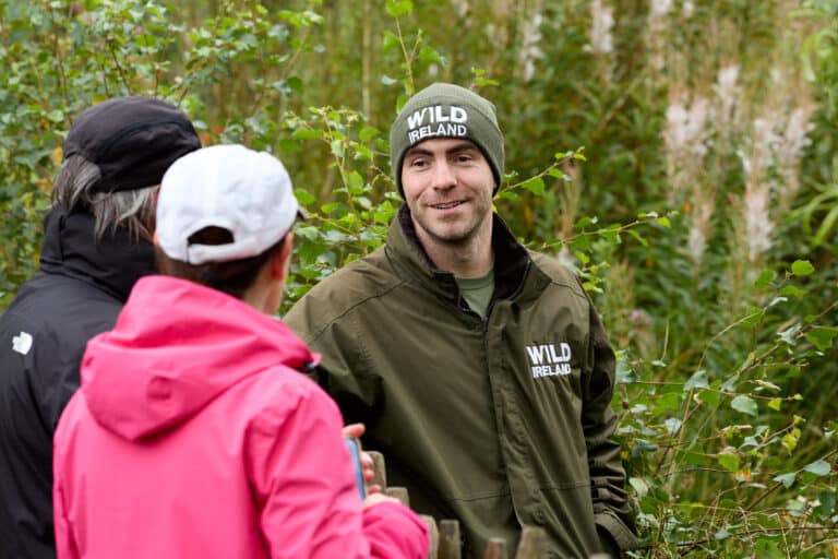 Wild Ireland - A person in a green jacket and beanie that read Wild Ireland stands outdoors, smiling and talking to two others. The setting is lush with greenery, and one person wears a pink jacket and white cap, while the other wears a black jacket and cap.