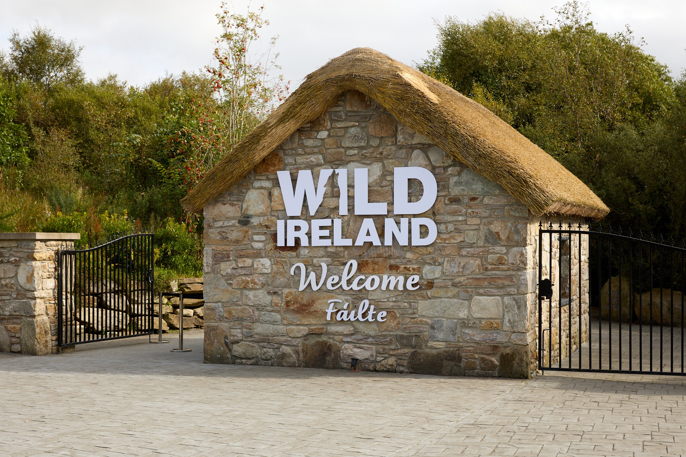 Wild Ireland - Stone entrance with a thatched roof displaying the words WILD IRELAND Welcome FƔilte. The building is surrounded by trees, and metal gates are open on either side.