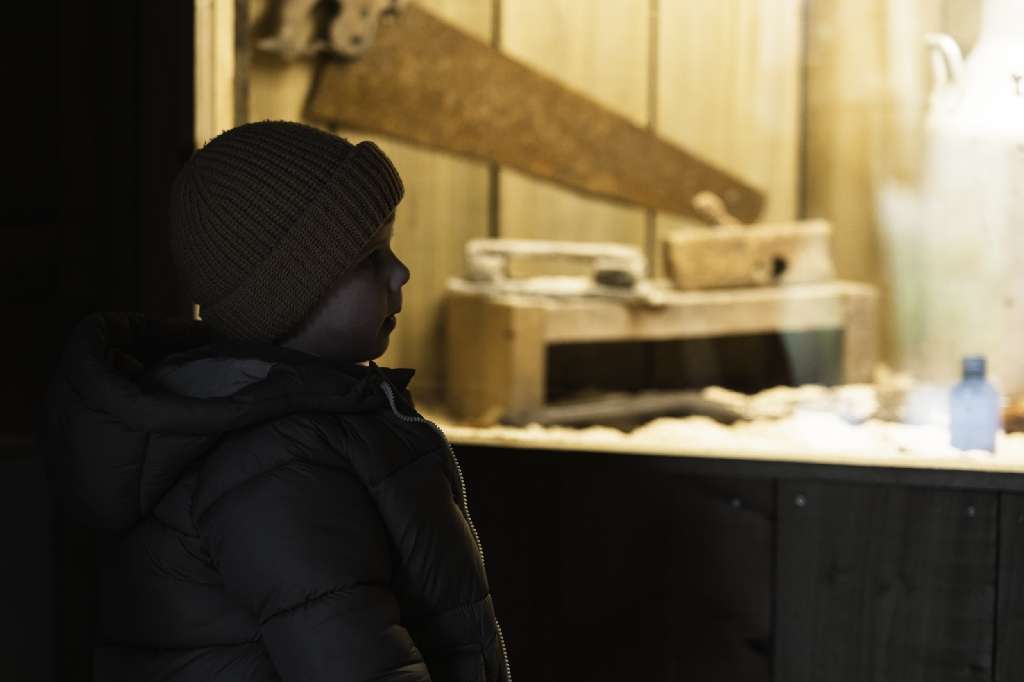 Wild Ireland - A child in a dark jacket and beanie observes a display case with wooden objects and a large saw. The scene is dimly lit, highlighting the objects inside the case.