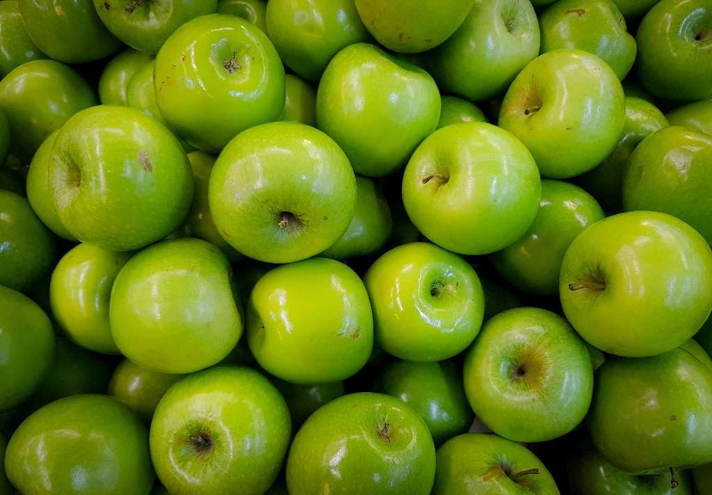 Wild Ireland - A pile of vibrant green apples with smooth, shiny skins is closely packed together. Some apples have subtle blemishes and stem ends are visible. The background is entirely filled with the apples, showcasing their uniform color and shape.