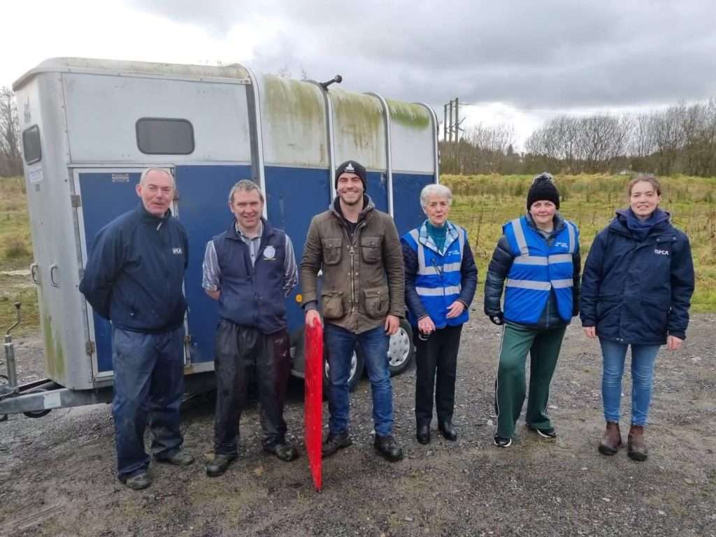 Wild Ireland - Six people stand in front of a horse trailer in an outdoor setting. Four wear blue jackets or vests, and one holds a red shield-like object. The sky is cloudy and grassy fields are in the background.