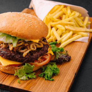 Wild Ireland - A sesame seed burger with lettuce, a beef patty, cheese, and crispy onions sits on a wooden board. A side of fries is wrapped in paper next to it. There is a garnish of mixed greens nearby.
