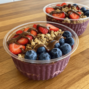 Wild Ireland - Two plastic bowls of acai smoothie topped with granola, sliced strawberries, whole blueberries, and a drizzle of chocolate sauce, placed on a wooden table.