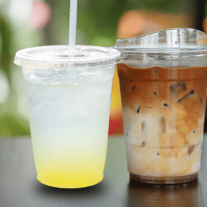 Wild Ireland - Two plastic cups with lids; one contains yellow lemonade with ice and a straw, the other has iced coffee with milk, ice cubes, and no straw. Both drinks are on a dark surface with a blurred green background.