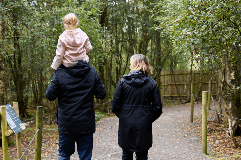 Wild Ireland - An adult carries a child on their shoulders while walking alongside another adult on a forest path. Both adults wear dark coats, and the child is in a light pink jacket. The path is lined with greenery and wooden posts.