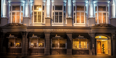 Wild Ireland - Exterior view of Bishop’s Gate Hotel, featuring illuminated upper windows and a warmly lit entrance. The facade includes several large windows and signage for Guinness, set against a dark-painted exterior.