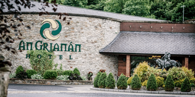 Wild Ireland - Stone building exterior with An Grianán Hotel signage. Entrance area surrounded by greenery and shrubs. A sculpture of a man on horseback is visible near the entrance. Forested area can be seen in the background.
