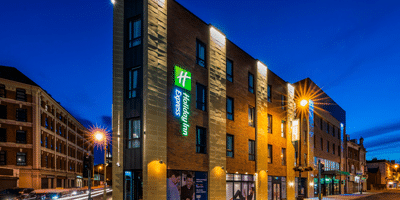Wild Ireland - A modern, multi-story Holiday Inn Express building lit up at night. The hotel features a prominent green and blue sign. Surrounding the hotel, streetlights illuminate the urban setting with a clear evening sky in the background.