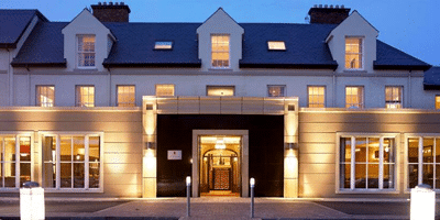 Wild Ireland - A large, modern two-story building with three dormer windows on the roof is illuminated at dusk. The facade features a central entrance flanked by tall windows. The building is set against a twilight sky.